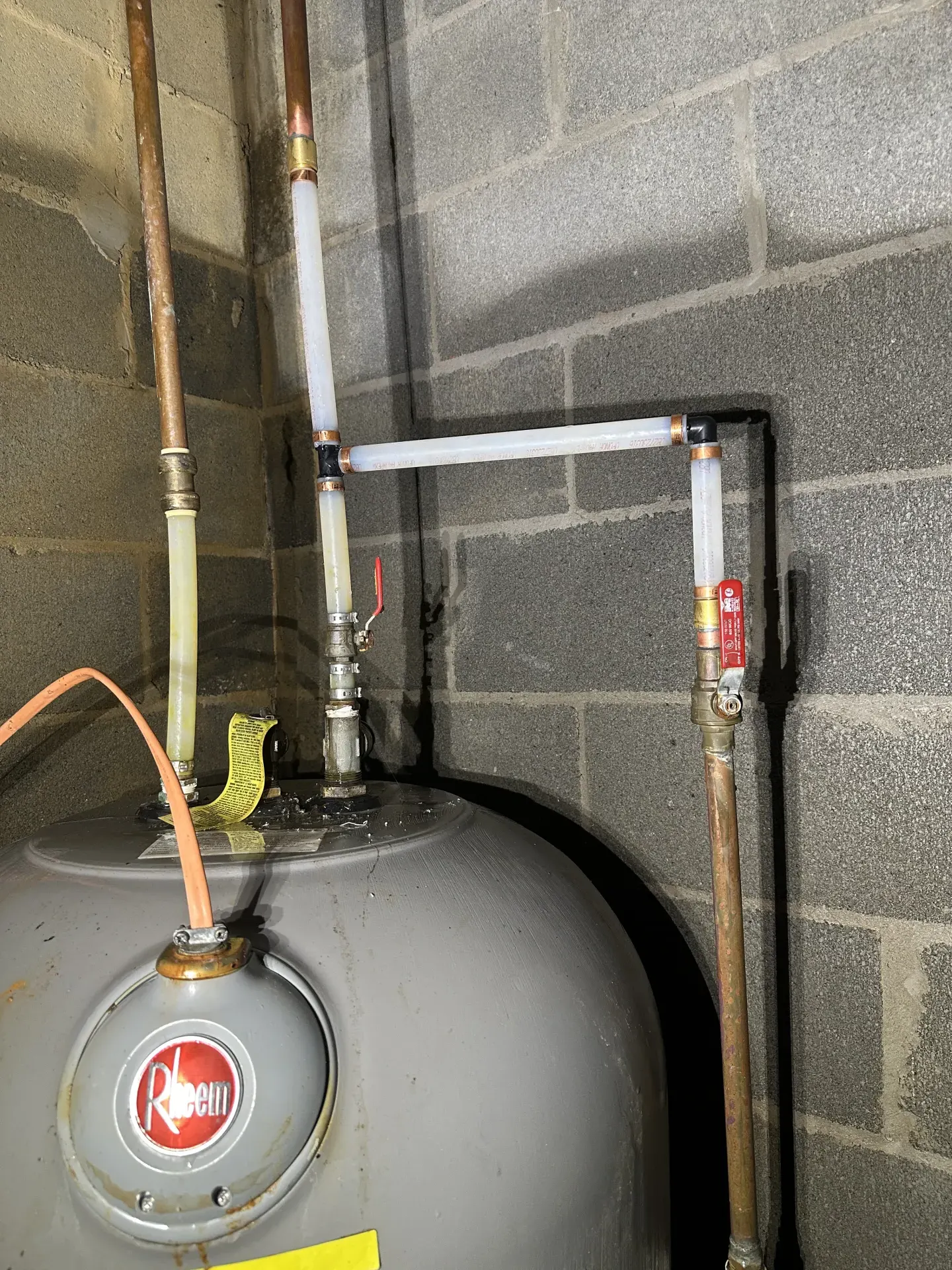 Close-up of a soldered copper elbow joint above a water heater tank, with a fine mist of water visibly spraying from a pinhole leak against the cinder block wall behind it.