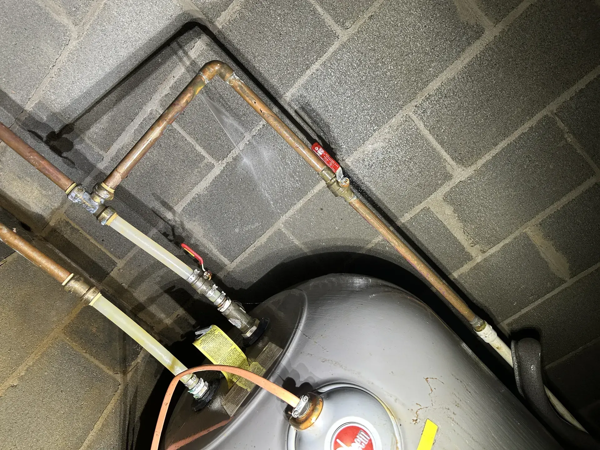 Close-up of a soldered copper elbow joint above a water heater tank, with a fine mist of water visibly spraying from a pinhole leak against the cinder block wall behind it